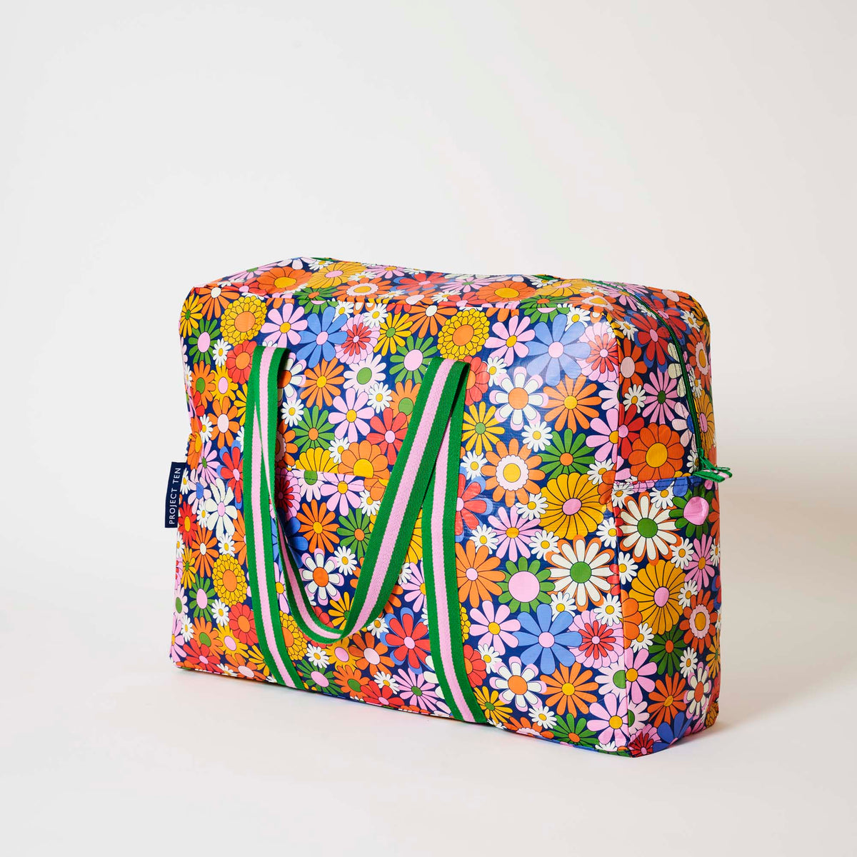 Our Coastal Floral Overnight Bag, a roomy duffle-style bag in a retro daisy print of orange, red, pink, yellow, blue and white flowers on a navy background, with two green and pink striped carry handles and a green zip.