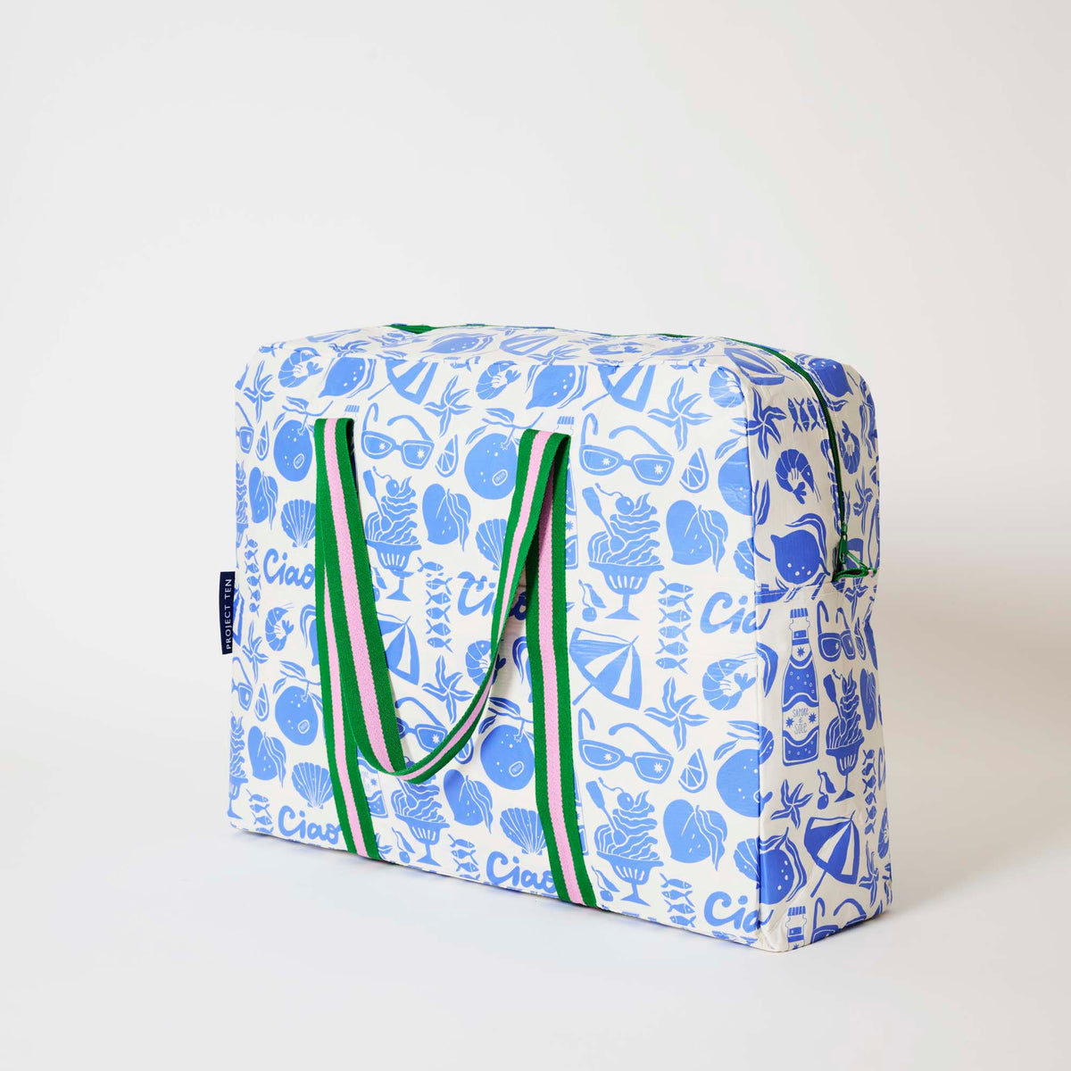 Our Feast Overnight Bag, a roomy duffle-style bag in an ecru base with a blue seaside print of sunglasses, ice cream sundaes, oranges, shells, sardines and "Ciao" lettering, with two green and pink striped carry handles and a green zip.