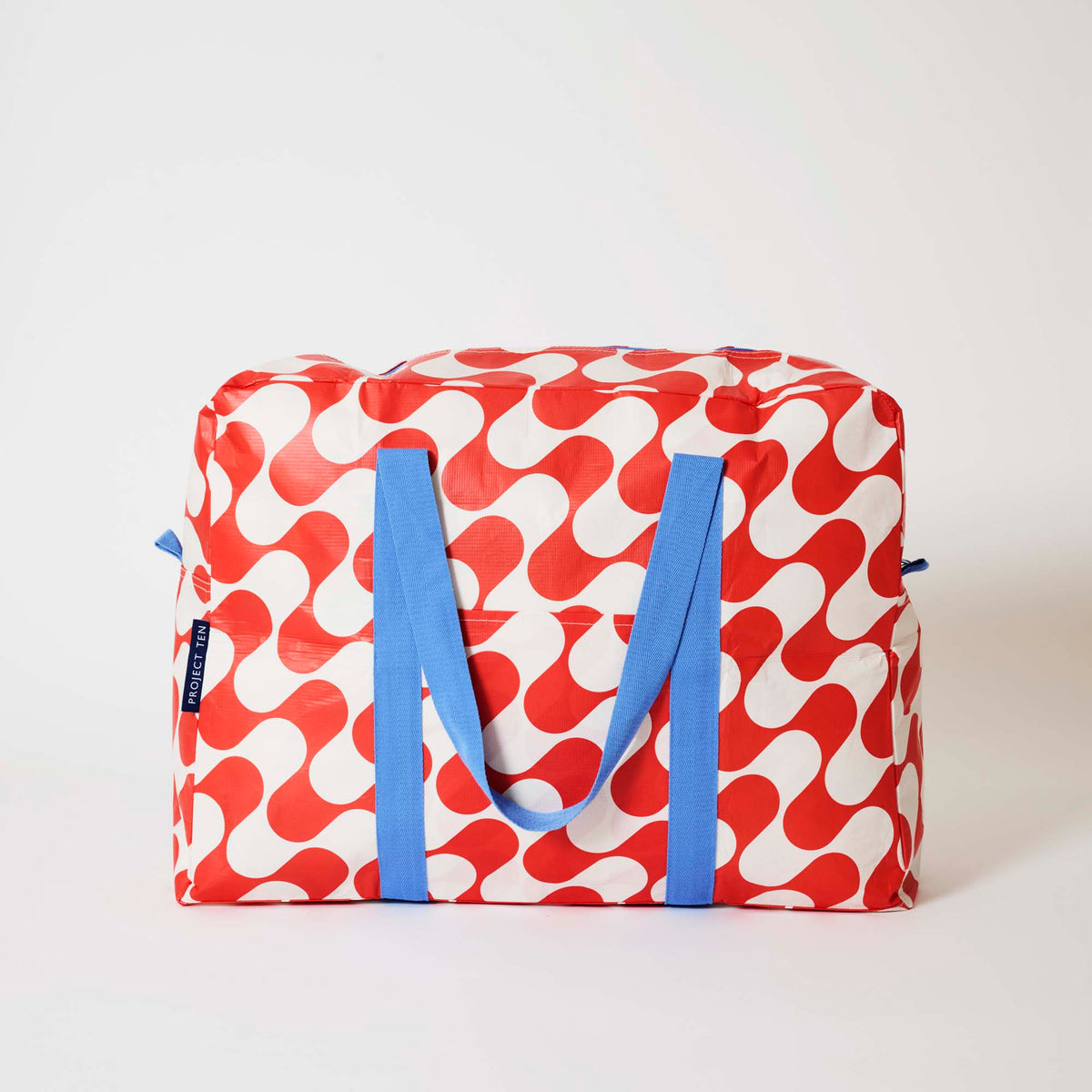 Our Red Ripple Overnight Bag, a roomy duffle-style bag in a bold geometric wave print of red and ecru, with two blue carry handles and a blue zip.