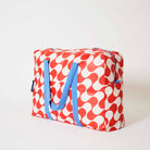 Our Red Ripple Overnight Bag, a roomy duffle-style bag in a bold geometric wave print of red and ecru, with two blue carry handles and a blue zip.