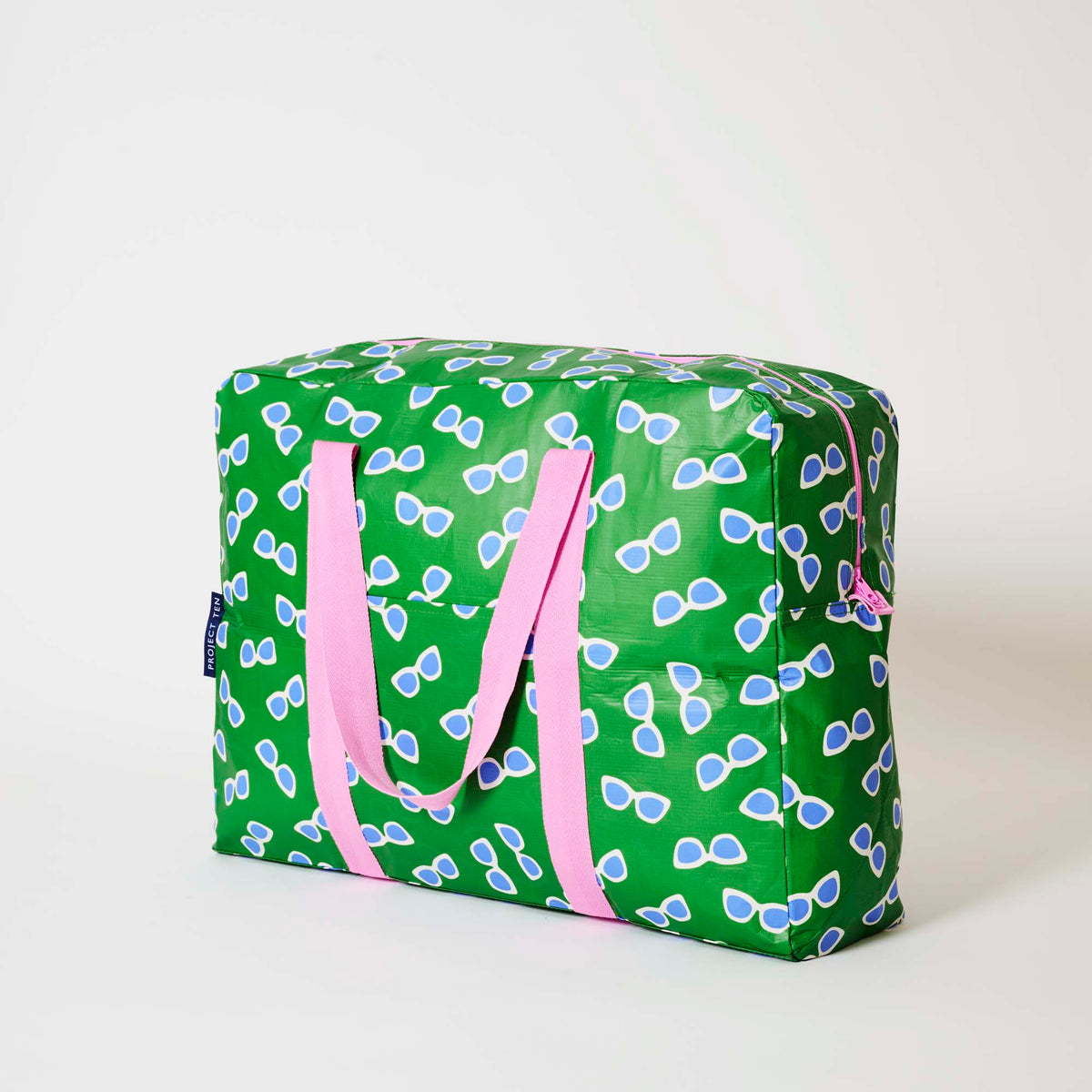 Our Sunglasses Overnight Bag, a roomy duffle-style bag in green with a scattered print of blue-lensed, white-framed sunglasses, two pink carry handles and a pink zip.