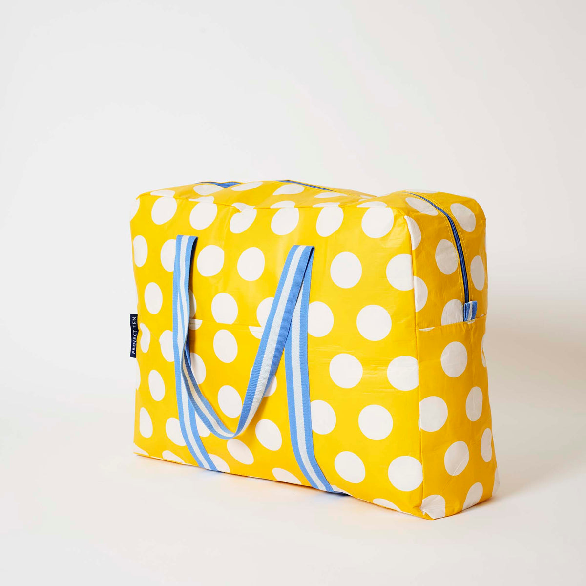 Our Yellow Polka Dot Overnight Bag, a roomy duffle-style bag in bright yellow with large ecru polka dots, two blue and ecru striped carry handles and a blue zip.