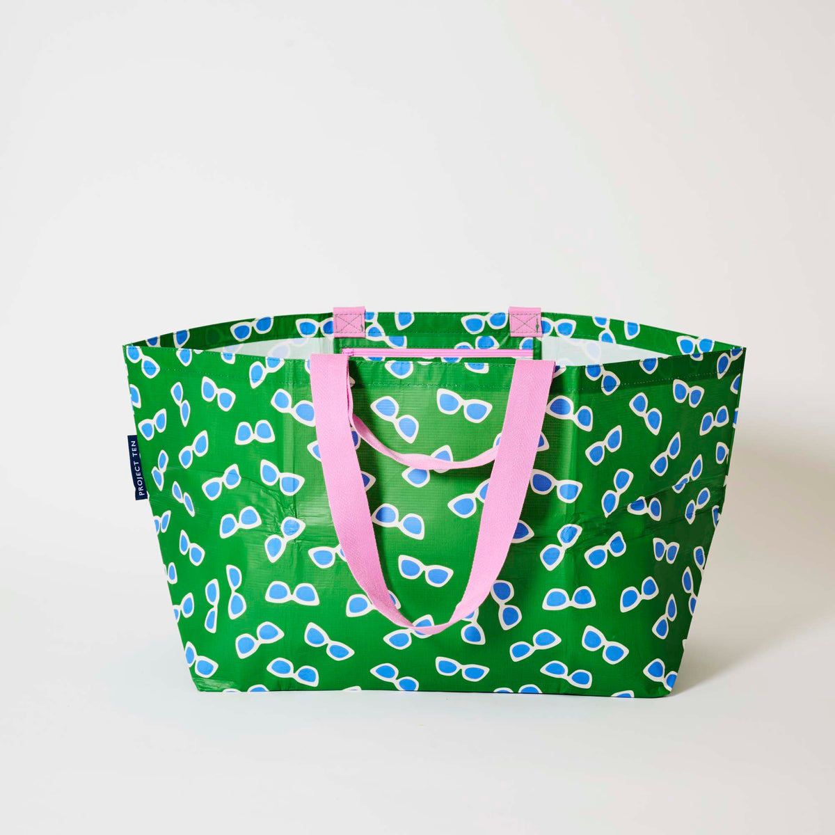 Our Sunglasses Oversized Tote in green with a scattered print of ecru framed sunglasses with blue lenses, and pink straps, shown open against a plain background.