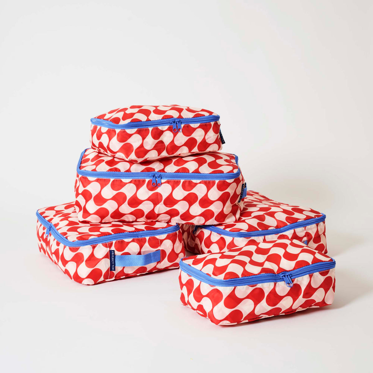 Our Red Ripple Packing Cubes, a set of five zip-up packing cubes in different sizes, in a bold geometric wave print of red and ecru, with blue zips and blue carry handles.