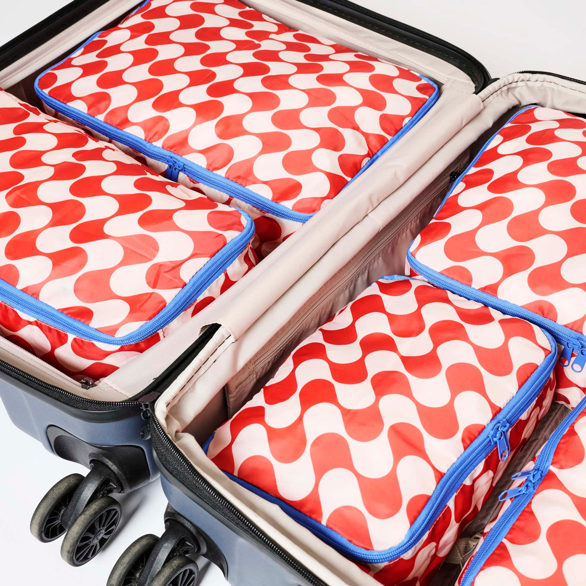 Our Red Ripple Packing Cubes, a set of five zip-up packing cubes in different sizes, in a bold geometric wave print of red and ecru, with blue zips and blue carry handles.