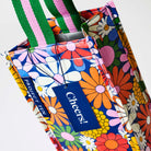 Our Coastal Floral Wine Bag, a tall narrow bottle bag in a retro daisy print of orange, red, pink, yellow and white flowers on a navy background, with a single green and pink striped carry handle.