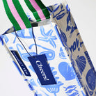 Our Feast Wine Bag, a tall narrow bottle bag in an ecru base with a blue seaside print of sunglasses, ice cream sundaes, beach umbrellas, starfish and sardines, with a single green and pink striped carry handle.
