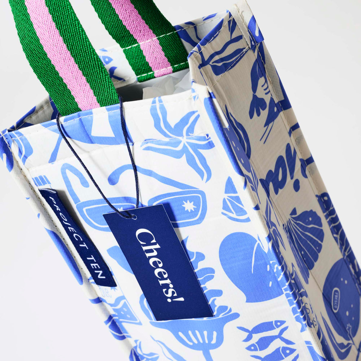 Our Feast Wine Bag, a tall narrow bottle bag in an ecru base with a blue seaside print of sunglasses, ice cream sundaes, beach umbrellas, starfish and sardines, with a single green and pink striped carry handle.