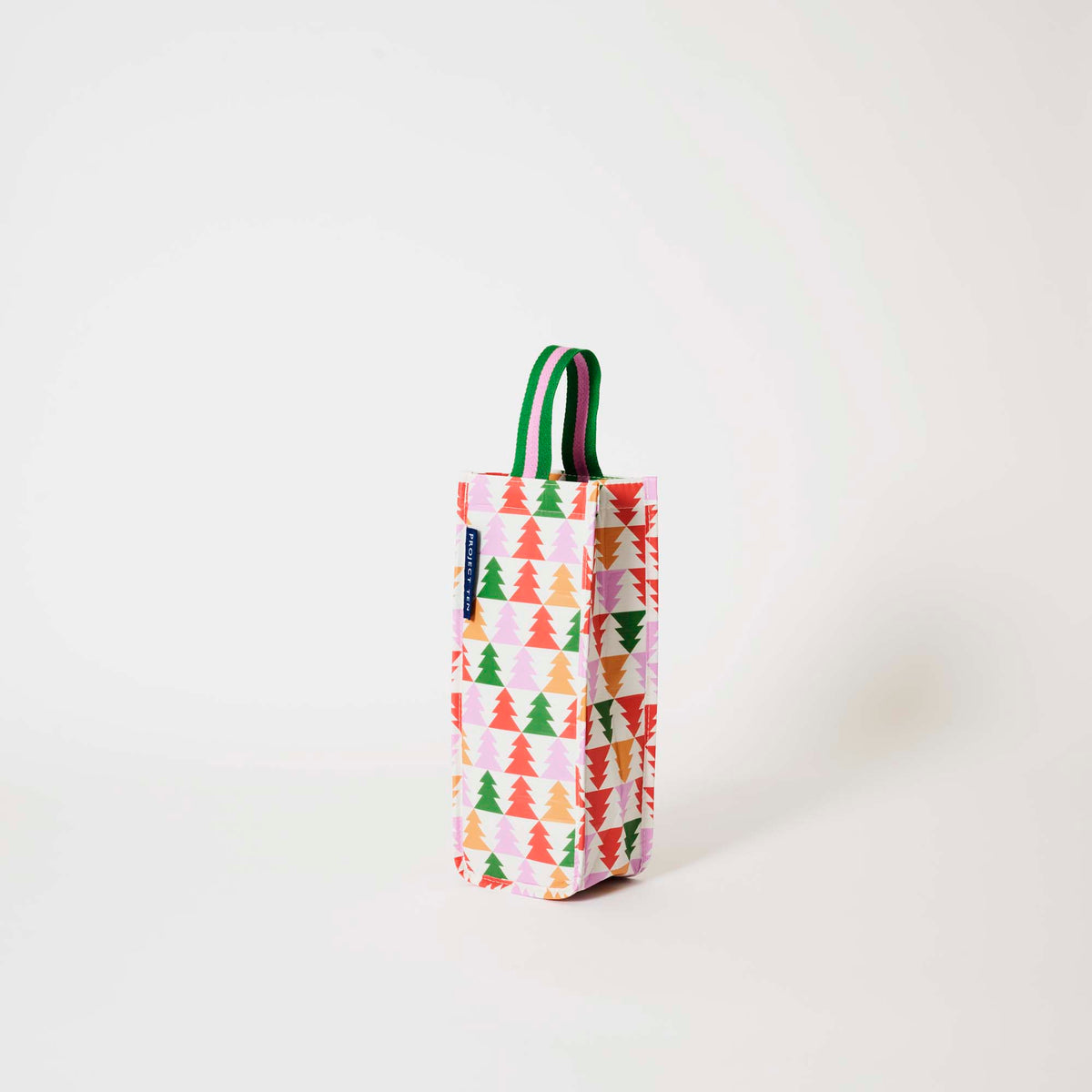 Our Holiday Cheers Wine Bag, a tall narrow bottle bag in an ecru base with a repeating geometric Christmas tree print in red, green, pink and orange, with a single green and pink striped carry handle.