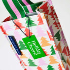 Our Holiday Cheers Wine Bag, a tall narrow bottle bag in an ecru base with a repeating geometric Christmas tree print in red, green, pink and orange, with a single green and pink striped carry handle.