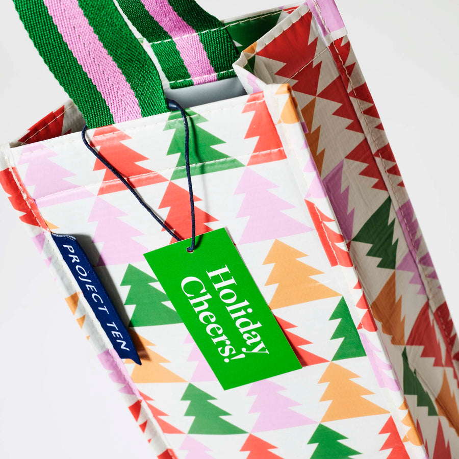 Our Holiday Cheers Wine Bag, a tall narrow bottle bag in an ecru base with a repeating geometric Christmas tree print in red, green, pink and orange, with a single green and pink striped carry handle.