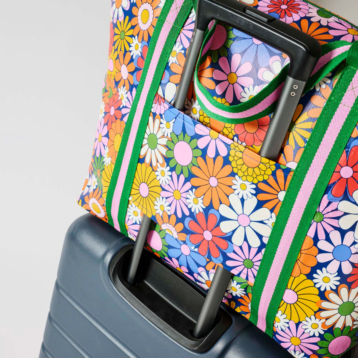 Our Coastal Floral Zip Medium Tote covered in bright retro daisies in orange, pink, yellow and blue on a navy background, with a full zip closure across the top and long green and pink striped shoulder straps attached to a suitcase.