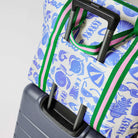 Our Feast Zip Medium Tote in a blue and ecru print of shells, gelato, soda bottles, sunglasses, beach umbrellas and the word "Ciao", with a full zip closure across the top and long green and pink striped shoulder straps attached to a suitcase.