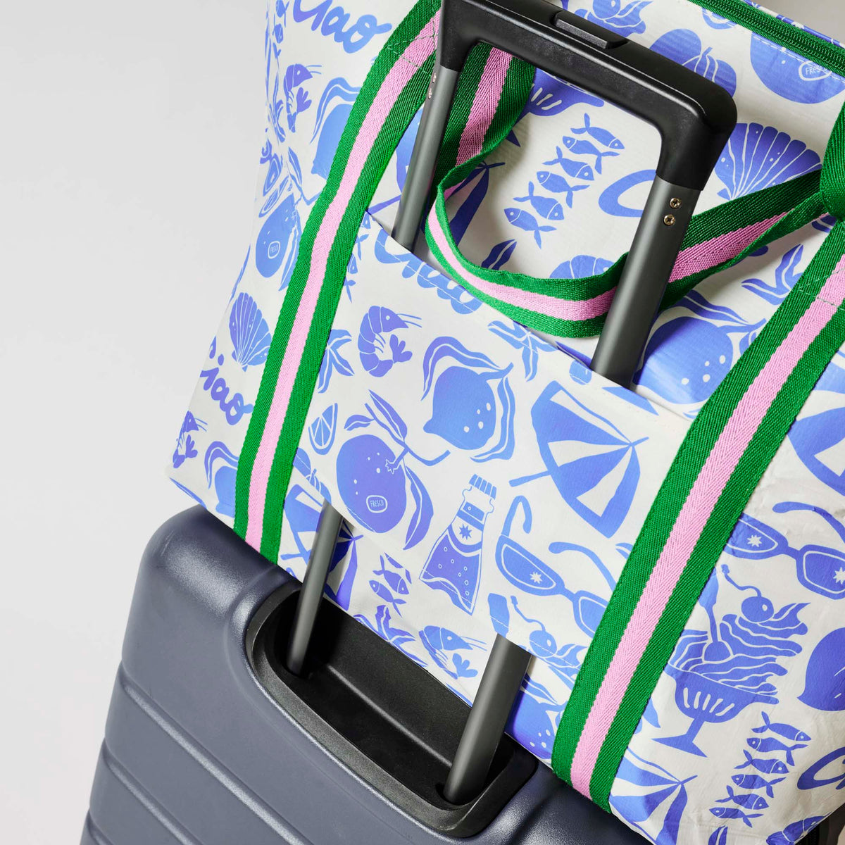 Our Feast Zip Medium Tote in a blue and ecru print of shells, gelato, soda bottles, sunglasses, beach umbrellas and the word "Ciao", with a full zip closure across the top and long green and pink striped shoulder straps attached to a suitcase.