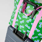 Our Sunglasses Zip Medium Tote in green with a scattered print of ecru framed sunglasses with blue lenses, with a full zip closure across the top and long pink shoulder straps attached to a suitcase.