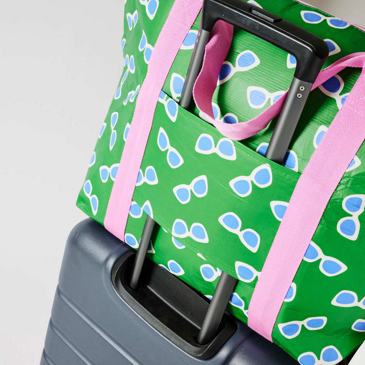 Our Sunglasses Zip Medium Tote in green with a scattered print of ecru framed sunglasses with blue lenses, with a full zip closure across the top and long pink shoulder straps attached to a suitcase.
