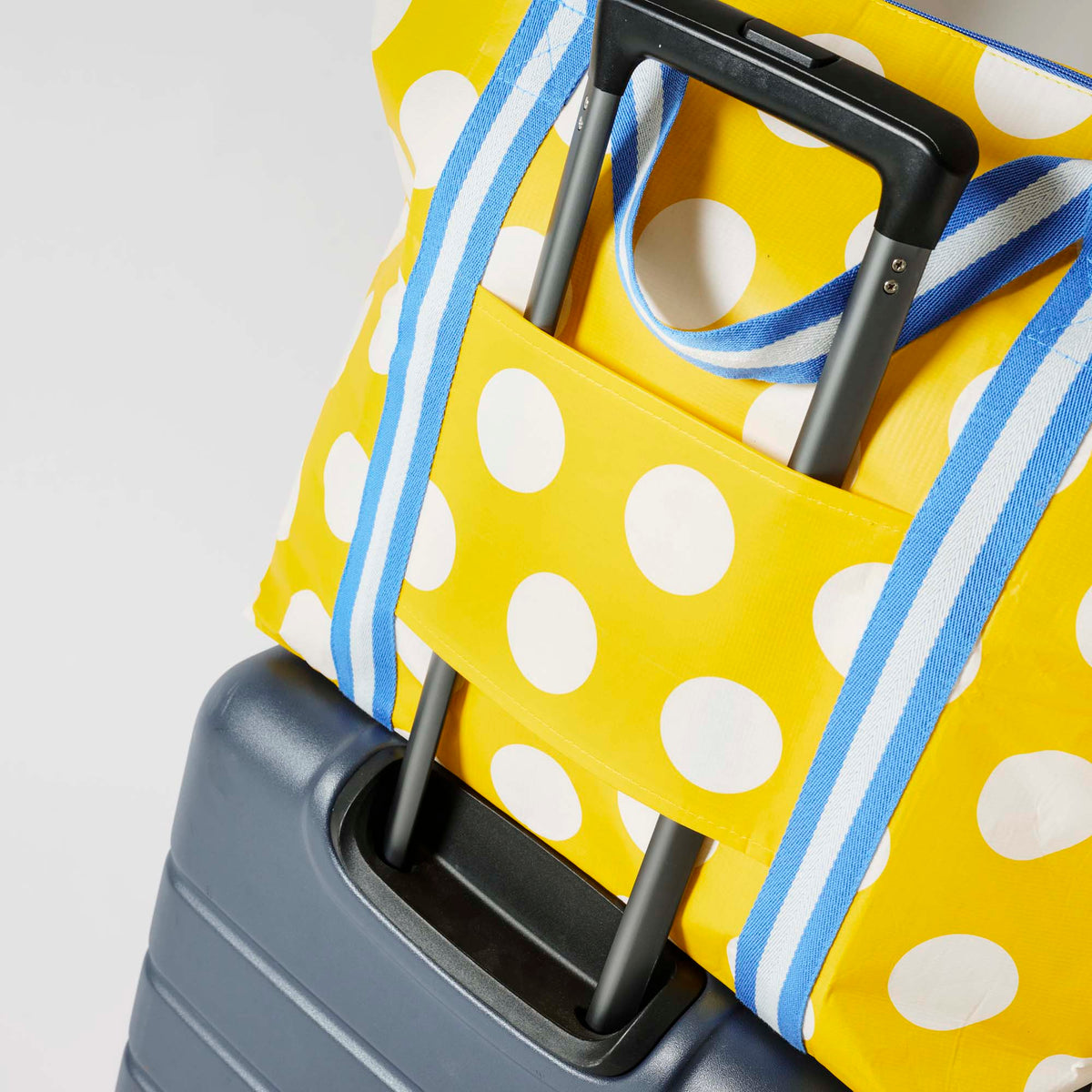 Our Yellow Polka Dot Zip Medium Tote in bright yellow with ecru polka dots, with a full zip closure across the top and long blue and ecru striped shoulder straps attached to a suitcase.